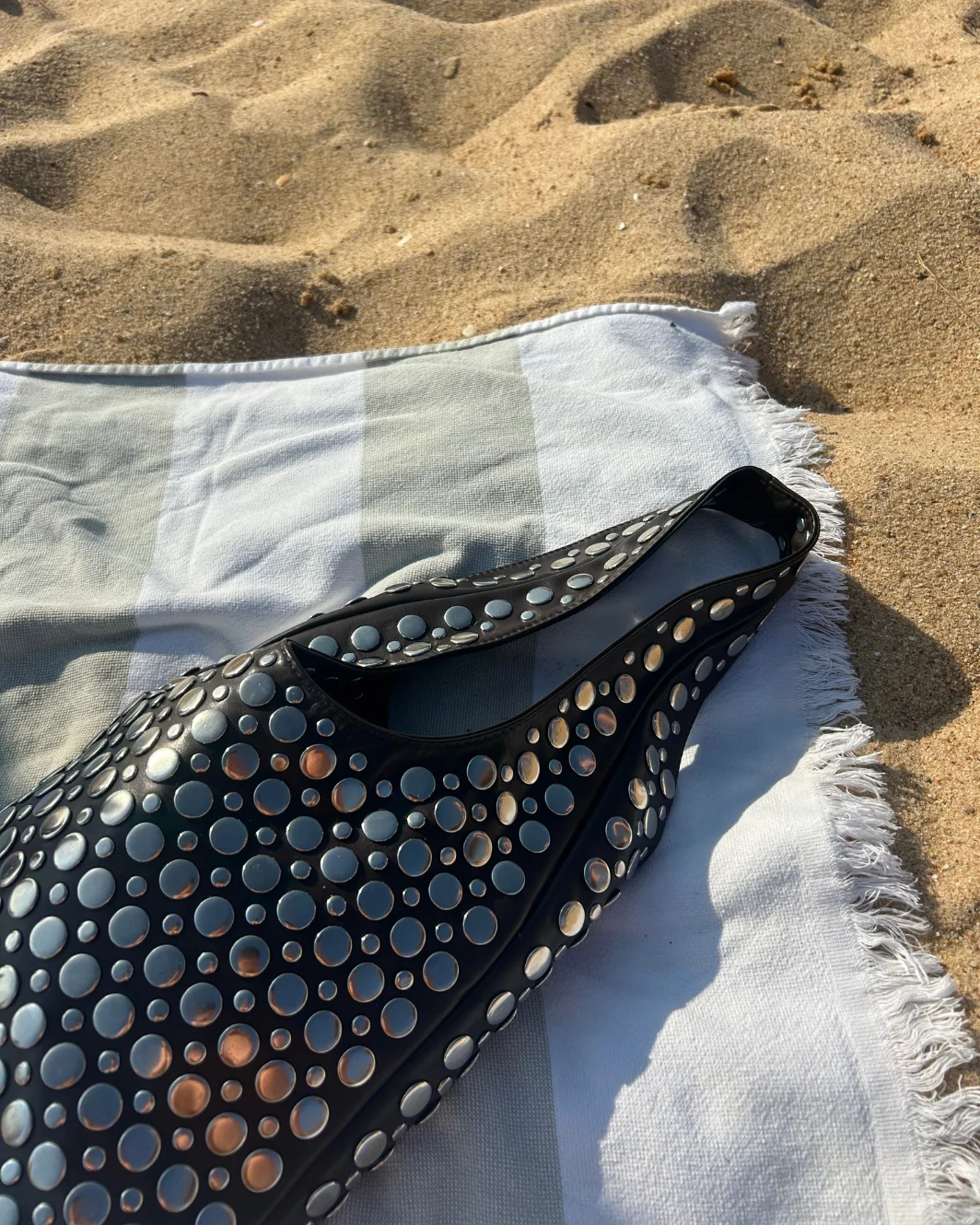 Black Christy studded shoulder bag resting on a towel on the sand at the beach.